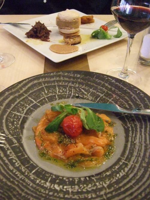 A plate of salmon tartare with a cherry tomato and greens, with a dessert and red wine in the background.