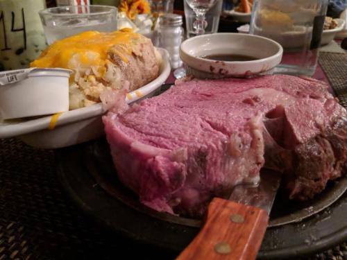 A large slice of prime rib on a sizzling plate, accompanied by a loaded baked potato and dipping sauce.