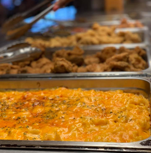 A buffet display featuring cheesy baked macaroni and cheese in the foreground, with fried chicken in the background.