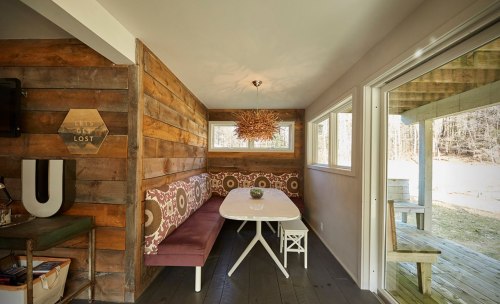 Cozy dining area with wooden walls, a white table, and a purple bench, featuring large windows and outdoor access.