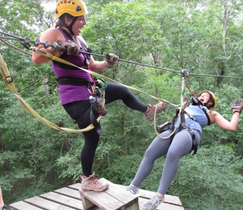 Two women in helmets and harnesses are on a zip line platform, one playfully kicking the other as they prepare to zip line.