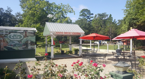 Outdoor café area with red umbrellas, tables, and a sign for "Abita Music Garden" surrounded by greenery and flowers.