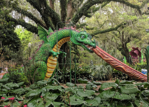 A colorful dragon sculpture with a long tongue, surrounded by lush greenery and trees in a whimsical garden setting.