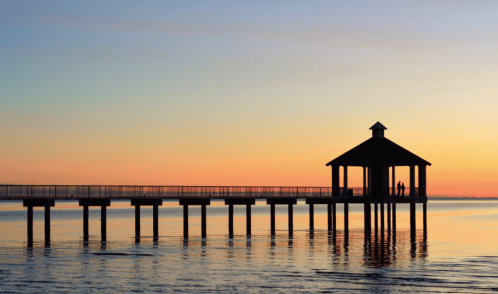 Silhouette of a pier with a gazebo at sunset, reflecting colors on calm water.
