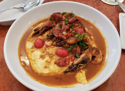 A bowl of shrimp and grits topped with tomatoes, green onions, and diced meat in a savory sauce.