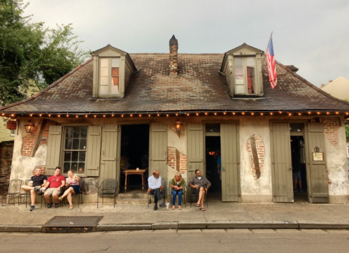A historic building with a sloped roof, featuring people sitting outside and an American flag hanging above the entrance.