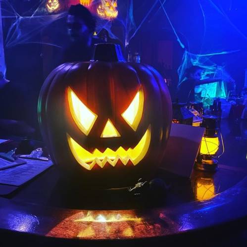 A glowing jack-o'-lantern with a menacing face sits on a bar, surrounded by Halloween decorations and dim lighting.