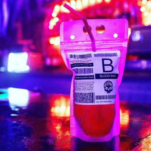 A vibrant pink drink in a blood bag-style container with a straw, set against a colorful, illuminated background.