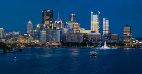 Pittsburgh skyline at dusk, featuring illuminated buildings and a river with a boat and fountain in the foreground.