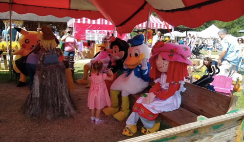 A child interacts with costumed characters, including a duck and a girl in a straw hat, under a colorful tent.