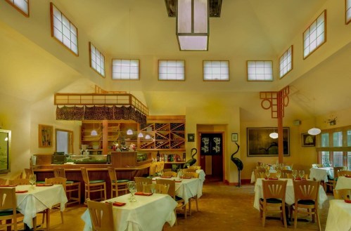 A spacious restaurant with yellow walls, elegant tables, and a bar area featuring wine racks and unique lighting.