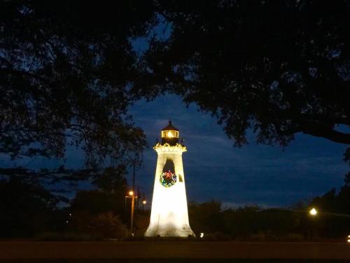 A brightly lit lighthouse adorned with a wreath, surrounded by trees at dusk.