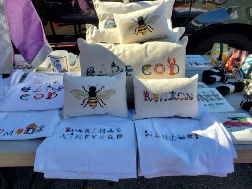 A display of decorative pillows and shirts featuring designs of bees and text like "Cape Cod" and "Martha's Vineyard."