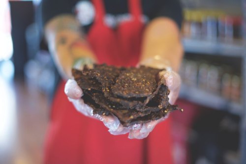 A person in a red apron holds a stack of dark, textured jerky in gloved hands, with shelves of jars in the background.