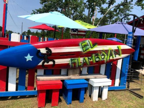 A colorful surfboard with a Texas flag design, featuring graffiti that reads "EL FLAMBOYAN," surrounded by picnic tables and umbrellas.