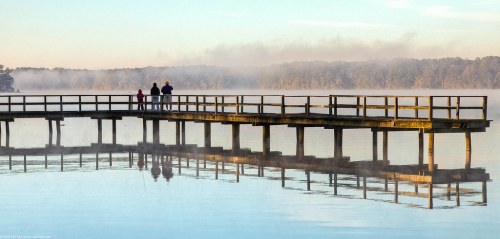 A serene lake scene with a wooden pier and three people silhouetted against a misty morning backdrop.