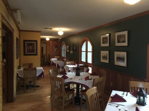 Cozy restaurant interior with wooden tables, white tablecloths, and framed artwork on the walls. Warm lighting.