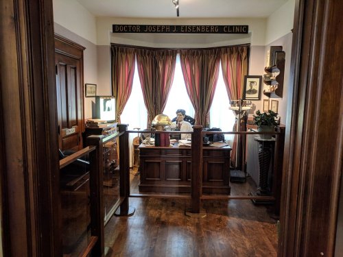 A vintage clinic office with wooden furniture, a globe, and a doctor seated at a desk, framed by curtains and historical photos.