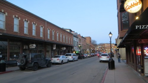 A quiet street lined with brick buildings, parked cars, and a restaurant sign, illuminated in the evening light.