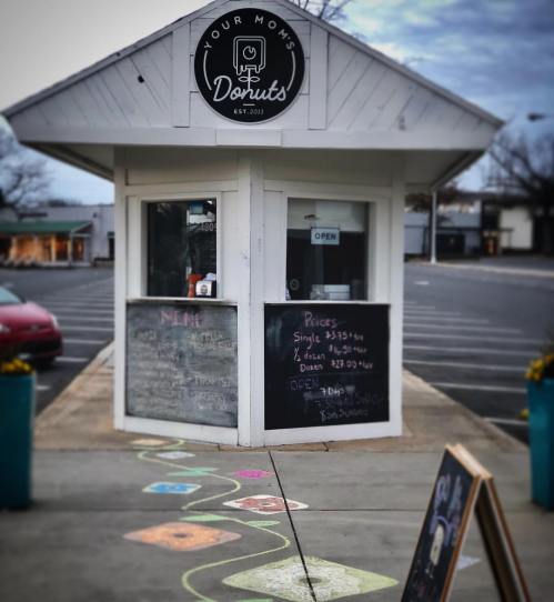A small donut shop with a chalk-drawn path leading to it, featuring a menu board and colorful flower pots.