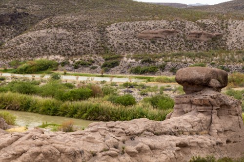 A rocky landscape features a large, flat-topped boulder beside a winding river and lush green vegetation.