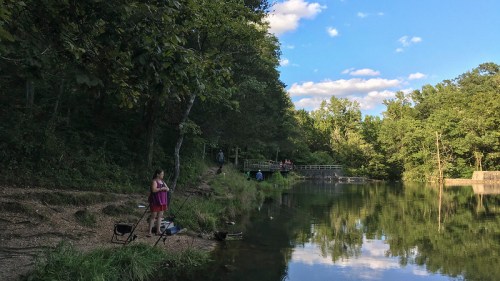 A serene lakeside scene with people fishing and enjoying nature under a blue sky with scattered clouds.