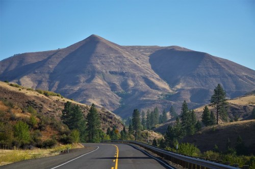 A winding road leads through a mountainous landscape with dry hills and scattered trees under a clear blue sky.