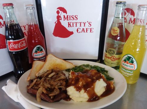 A plate of food with sliced meat, mashed potatoes, green beans, and drinks, set against Miss Kitty's Cafe signage.