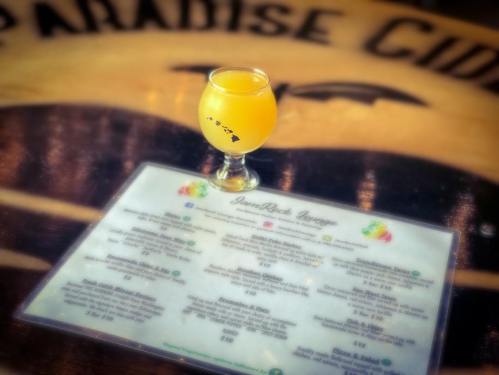 A glass of orange drink on a wooden table with a menu featuring various beverages and prices.
