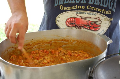 A person stirs a large pot of crawfish stew, wearing a shirt featuring a lobster graphic.