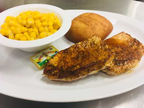 A plate with two seasoned pork chops, a serving of corn, a roll, and a butter packet.