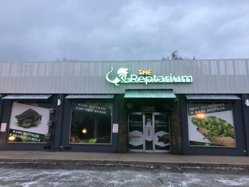 Exterior of The Reptarium, featuring large windows with images of reptiles and a neon sign, under a cloudy sky.