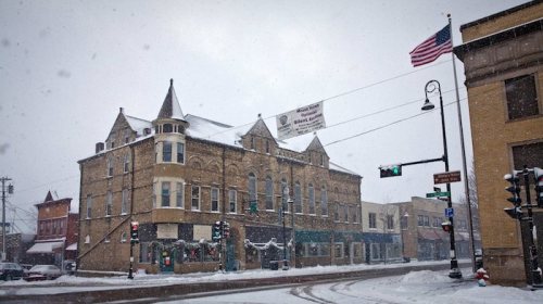 A snowy street scene featuring a historic building, traffic lights, and an American flag in a small town.