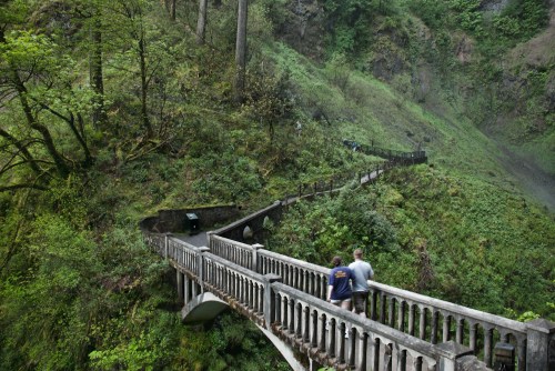 Two people walk along a winding stone bridge surrounded by lush greenery and steep hills.