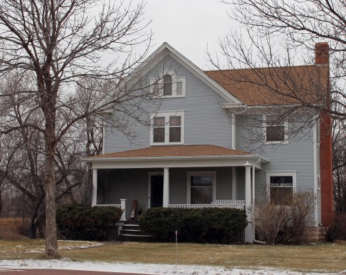 A large, light blue house with a porch, surrounded by bare trees and a snowy landscape.