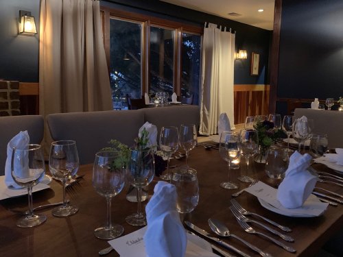 A dimly lit restaurant table set with glassware and folded napkins, overlooking a window with curtains drawn.
