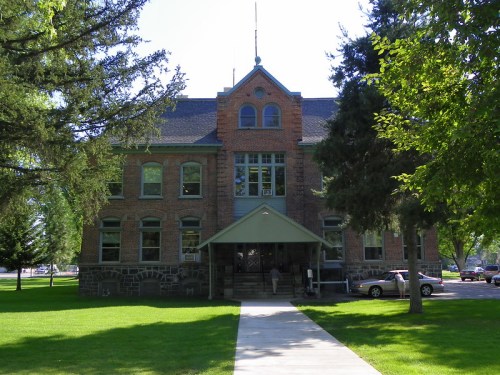 Historic brick building with a green awning, surrounded by trees and a grassy area, featuring large windows and a central entrance.