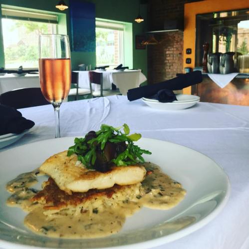 A plate of fish with sauce and greens, accompanied by a glass of rosé wine, set on a dining table.