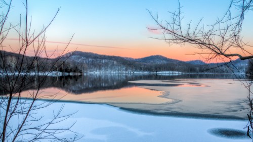 A serene winter landscape featuring a frozen lake, snow-covered banks, and soft pastel colors in the sky at sunset.
