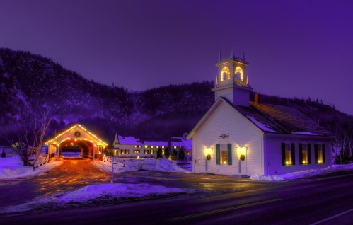 A snowy scene at dusk featuring a lit church and a covered bridge, adorned with festive decorations.