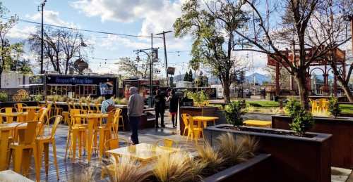 Outdoor dining area with yellow tables, trees, and food trucks under a blue sky with scattered clouds.