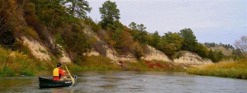 A person paddles a canoe on a calm river, surrounded by lush greenery and rocky cliffs in the background.