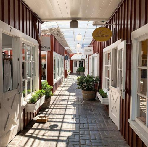 A sunlit corridor lined with shops, featuring red walls, white doors, and potted plants, creating a charming atmosphere.
