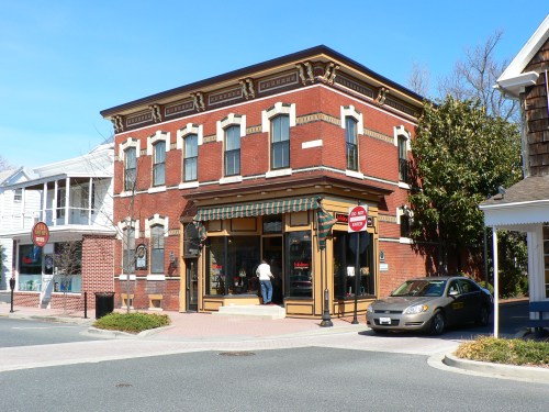 A brick building with a storefront, featuring large windows and a green awning, set in a quaint town square.