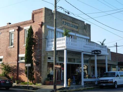 A charming brick building with a white balcony, featuring a café and shops, set against a clear blue sky.