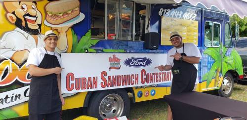 Two chefs in aprons stand beside a sign for a Cuban sandwich contest in front of a colorful food truck.