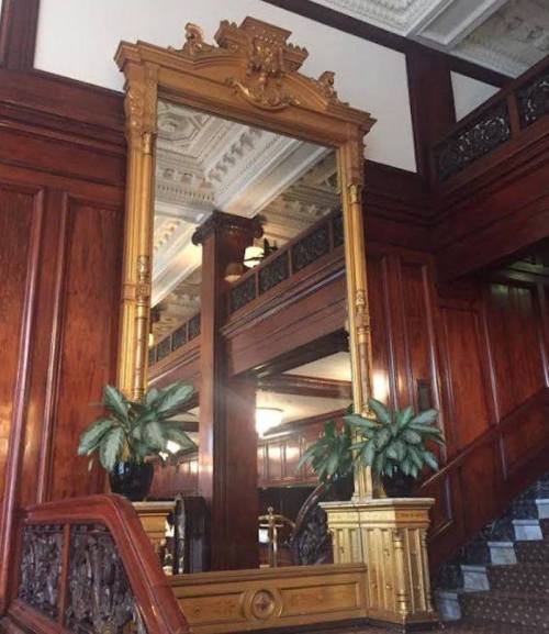 A large ornate gold mirror framed by decorative wood, surrounded by potted plants in a grand interior setting.