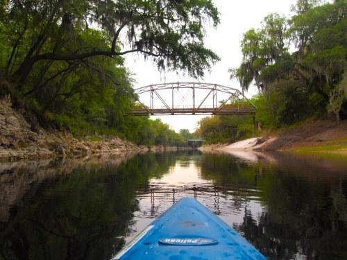 A kayak view of a calm river with lush greenery and a bridge in the background, reflecting on the water's surface.