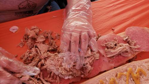 A gloved hand prepares a sandwich with shredded meat on a large roll, set on an orange tablecloth.