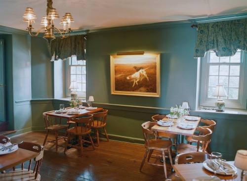 Cozy dining room with wooden tables, chairs, and a landscape painting on the wall, illuminated by soft lighting.
